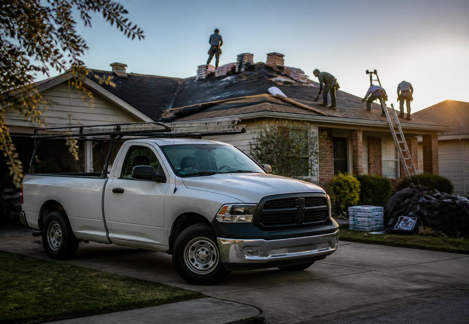 Placeholder: a contractor pickup truck in a driveway with a roofing crew working on the house behind it